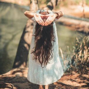 A woman with long hair performing a yoga pose by a tranquil river in a sunny outdoor setting.