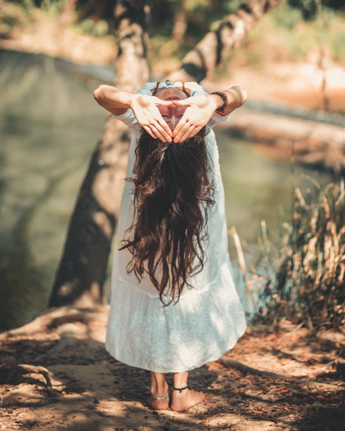 gallery-04 A woman with long hair performing a yoga pose by a tranquil river in a sunny outdoor setting.